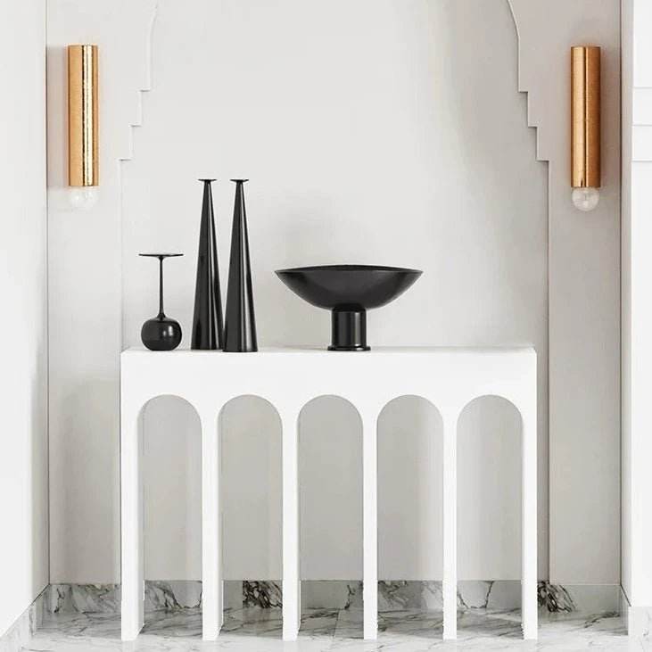 A white console table with a minimalist design, featuring arch-shaped cutouts at the bottom. It is placed against a wall with wall-mounted lamps on either side, and decorative objects are placed on top of the table.