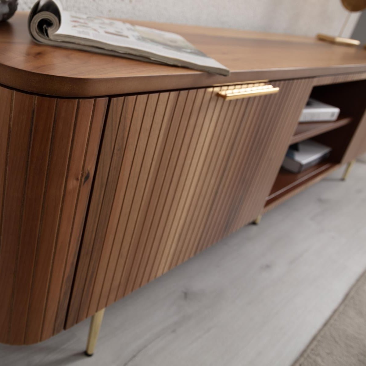 Asta Wooden TV Unit 200cm. Angled close-up of the media console in a living room setting, emphasizing the warm brown walnut wood with its diagonal slatted door design on the cabinet. The image shows the brass handle and thin brass legs, along with a magazine resting on the rounded top, demonstrating its functional, stylish storage for living areas.