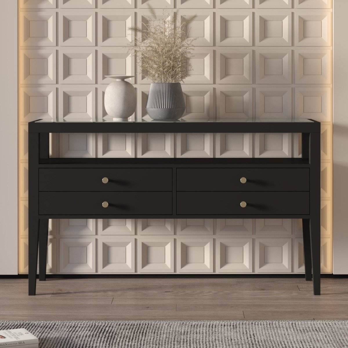 A sleek modern console table in black with brushed gold handles, featuring a tempered clear glass top, placed against a wall with a herringbone pattern. The table has four drawers and is positioned on a grey area rug with a white flower vase on top.