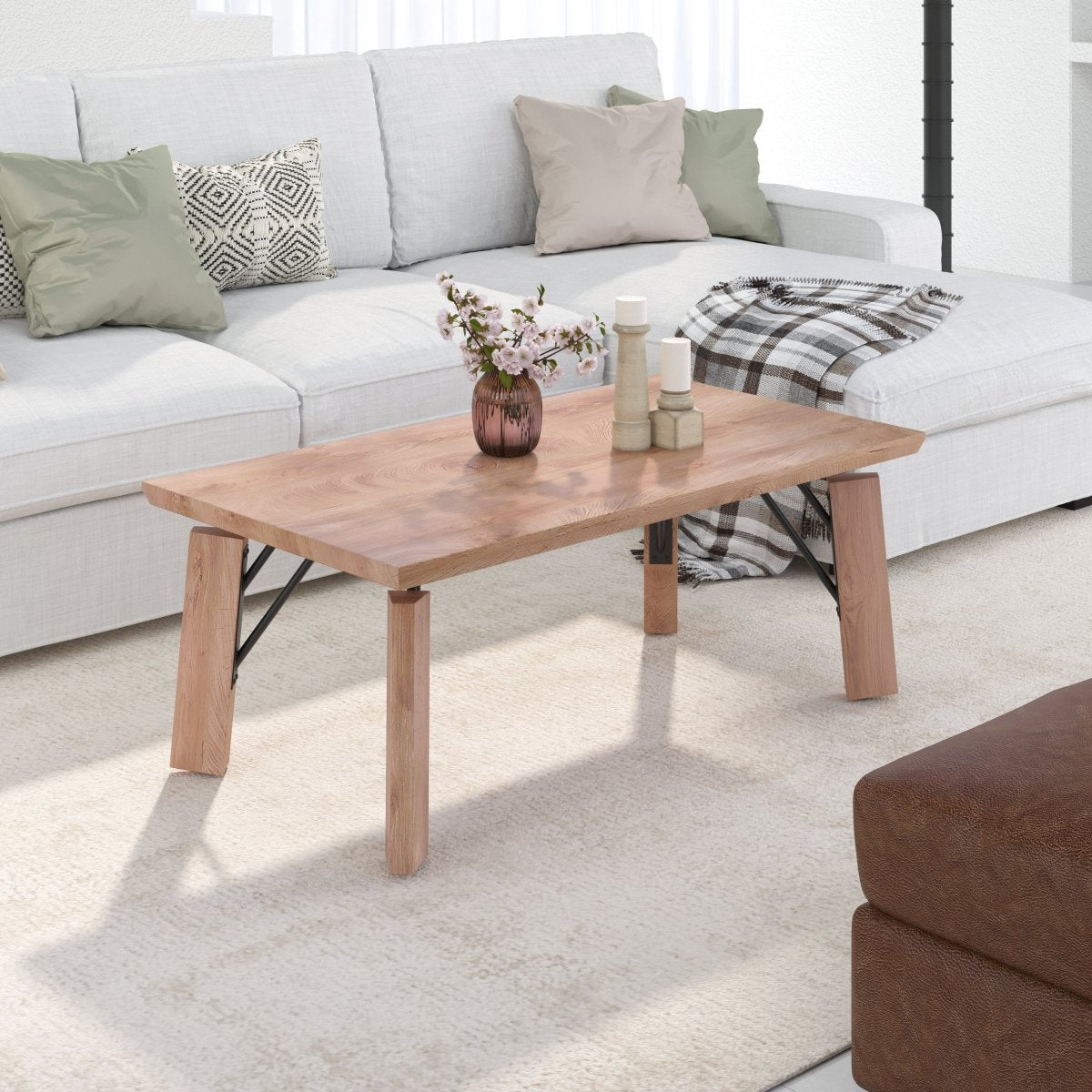 A rectangular wooden coffee table with thick legs and black metal support braces, styled in a bright living room next to a white sectional sofa.