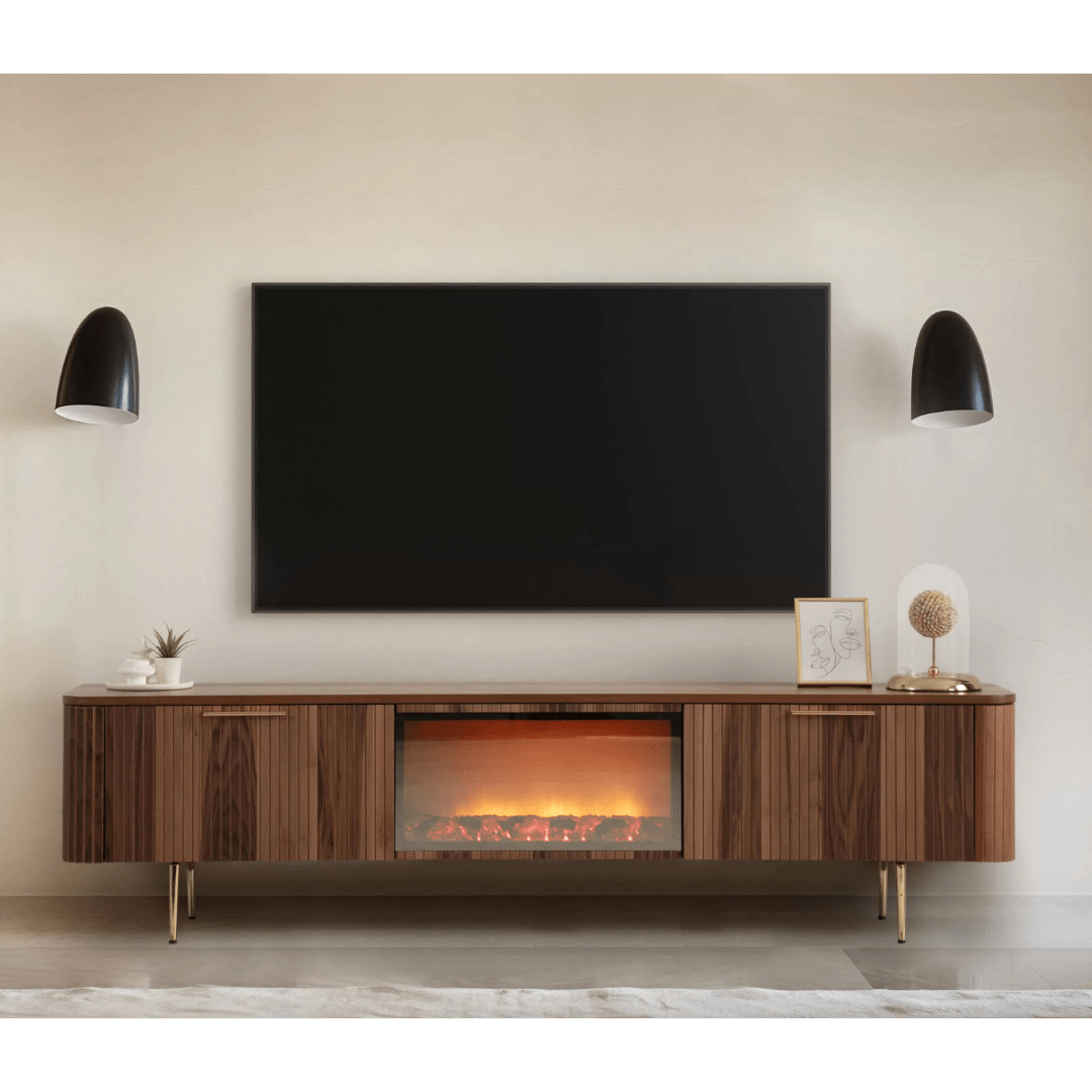 Asta Wooden Fire TV Unit 200cm. Long, mid-century modern style TV stand made of warm brown walnut wood, featuring two slatted storage cabinets on the sides and an integrated central electric fireplace insert with glowing logs. The unit is supported by slender brass legs and is shown beneath a large wall-mounted TV in a stylish living room setup, providing both storage and ambient.