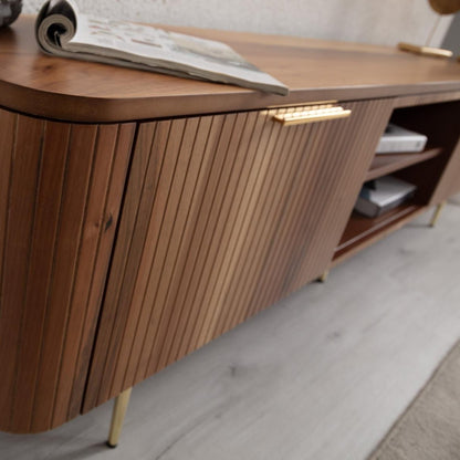 Asta Wooden TV Unit 200cm. Angled close-up of the media console in a living room setting, emphasizing the warm brown walnut wood with its diagonal slatted door design on the cabinet. The image shows the brass handle and thin brass legs, along with a magazine resting on the rounded top, demonstrating its functional, stylish storage for living areas.