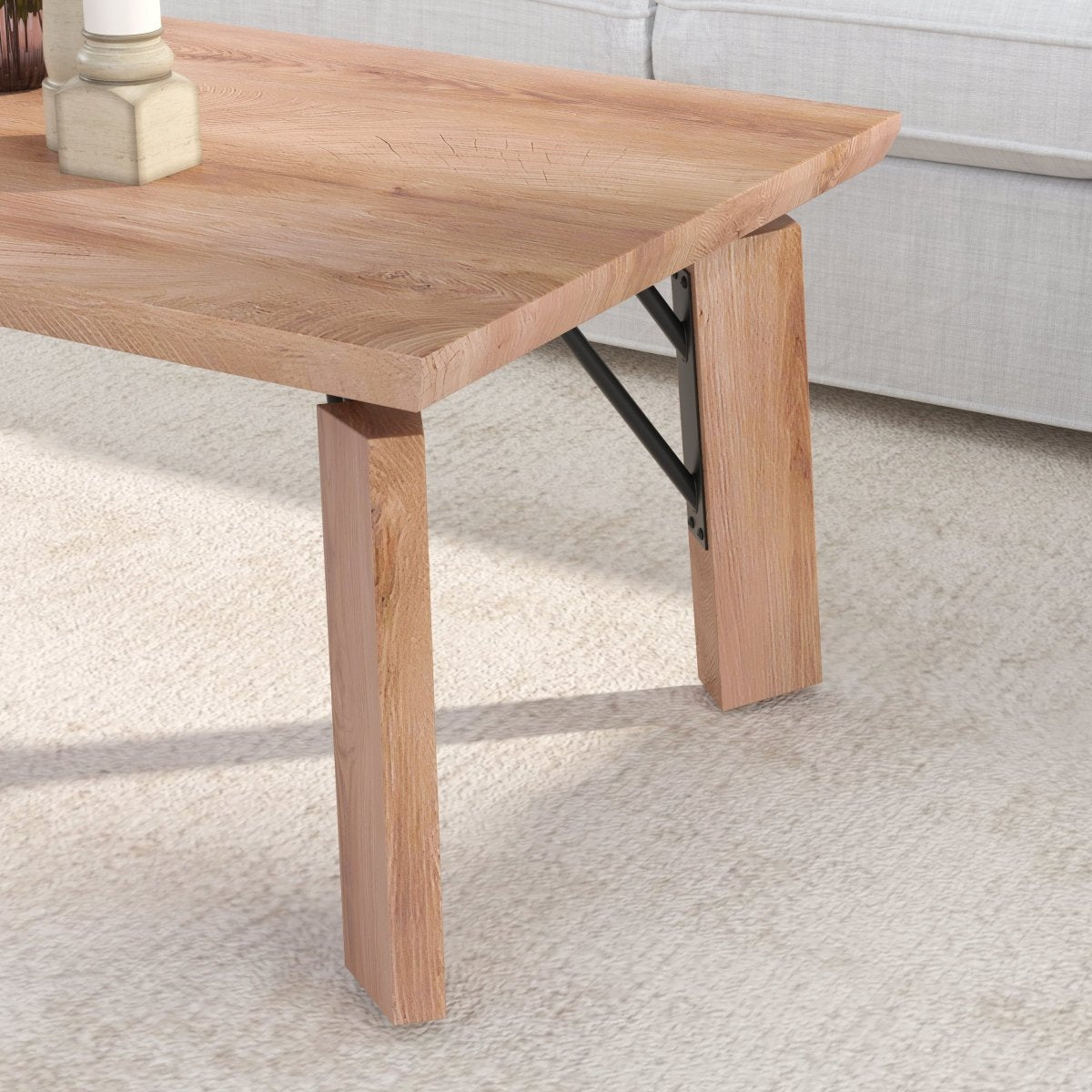 A close-up of a light oak wood coffee table with blocky legs and black metal support brackets, set on a cream carpet next to a light gray sofa.