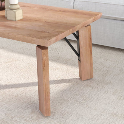 A close-up of a light oak wood coffee table with blocky legs and black metal support brackets, set on a cream carpet next to a light gray sofa.
