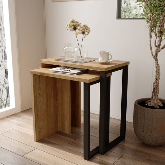 A small nesting side table set with a light oak wood top and black metal frame legs, styled in a bright room with a mug, glass vase, and dried flowers on top.