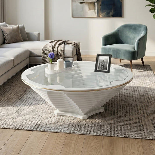 A modern round coffee table with a ribbed white pedestal base and a clear glass top, styled in a contemporary living room next to a gray sofa and a teal armchair.