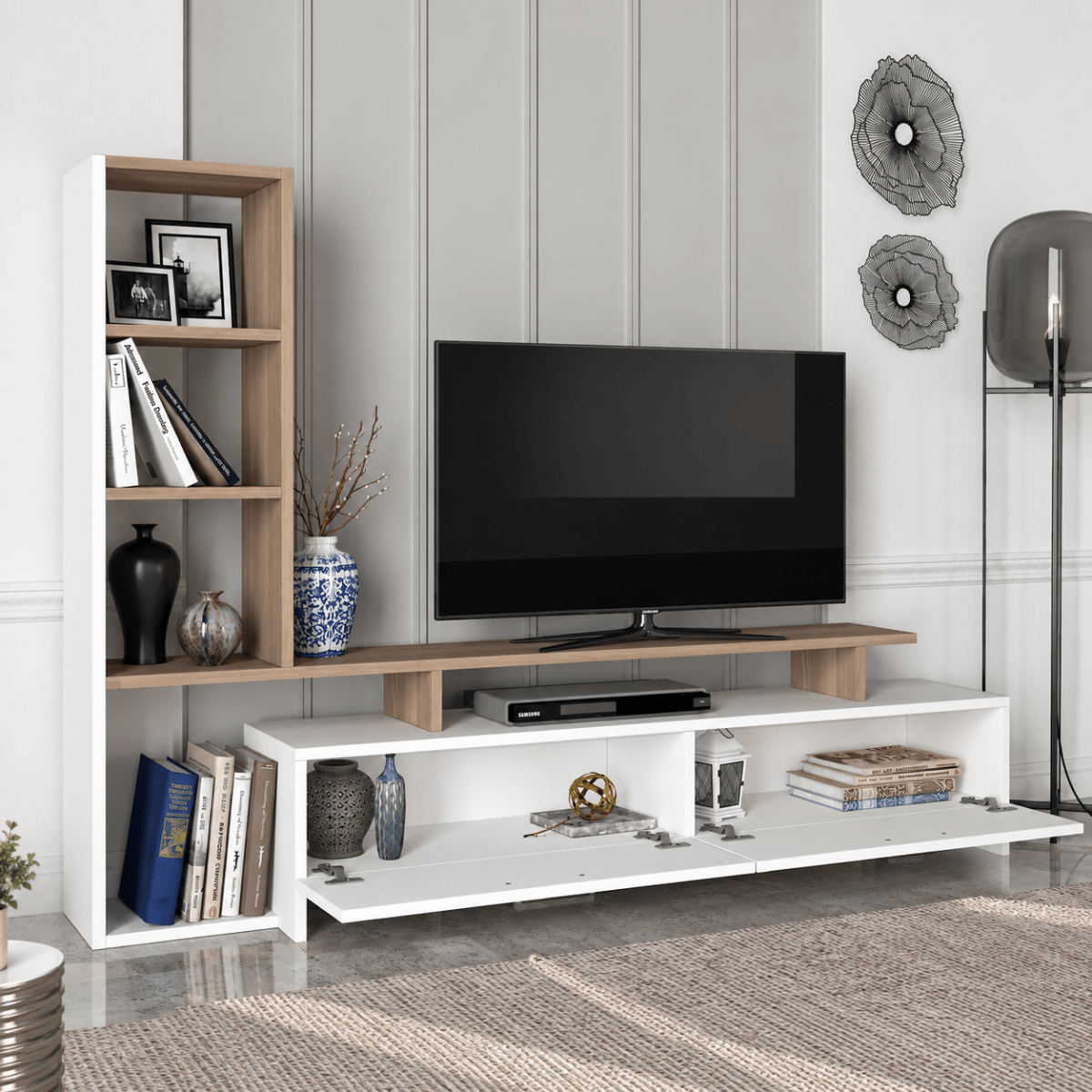 A modern white and light wood TV stand console with open shelving, styled in a living room setting with a flat-screen television, decorative vases, books, and a floor lamp.