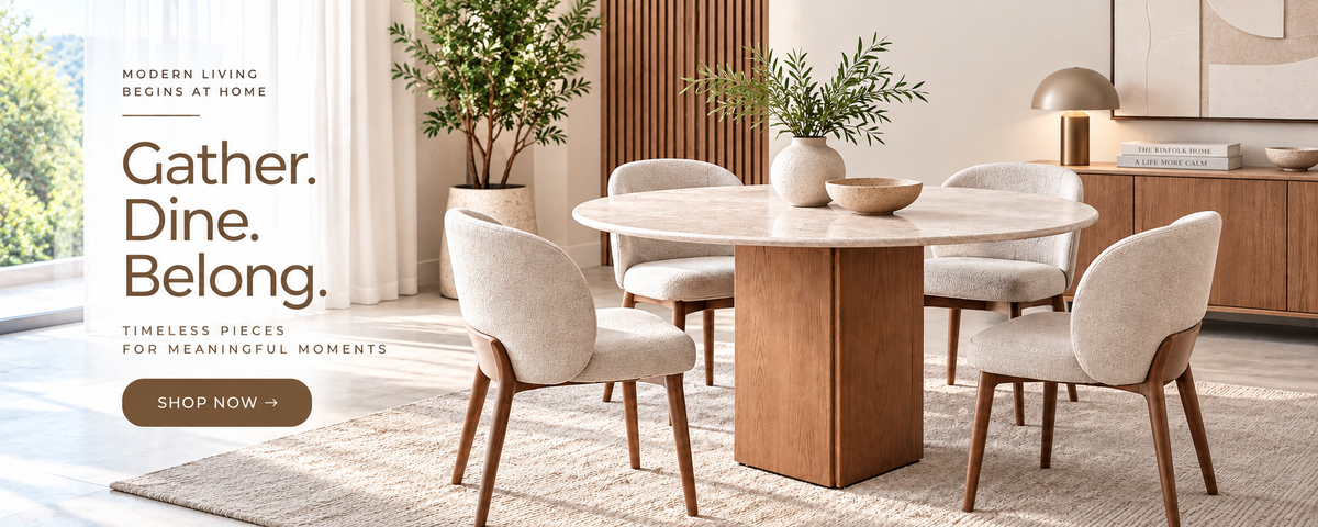A modern dining room scene showing a round light stone-top dining table with a substantial wooden pedestal base, surrounded by six upholstered dining chairs with light beige fabric and medium wood legs. The room has warm wood cabinetry, a sideboard, a potted plant, and a large window on the left.