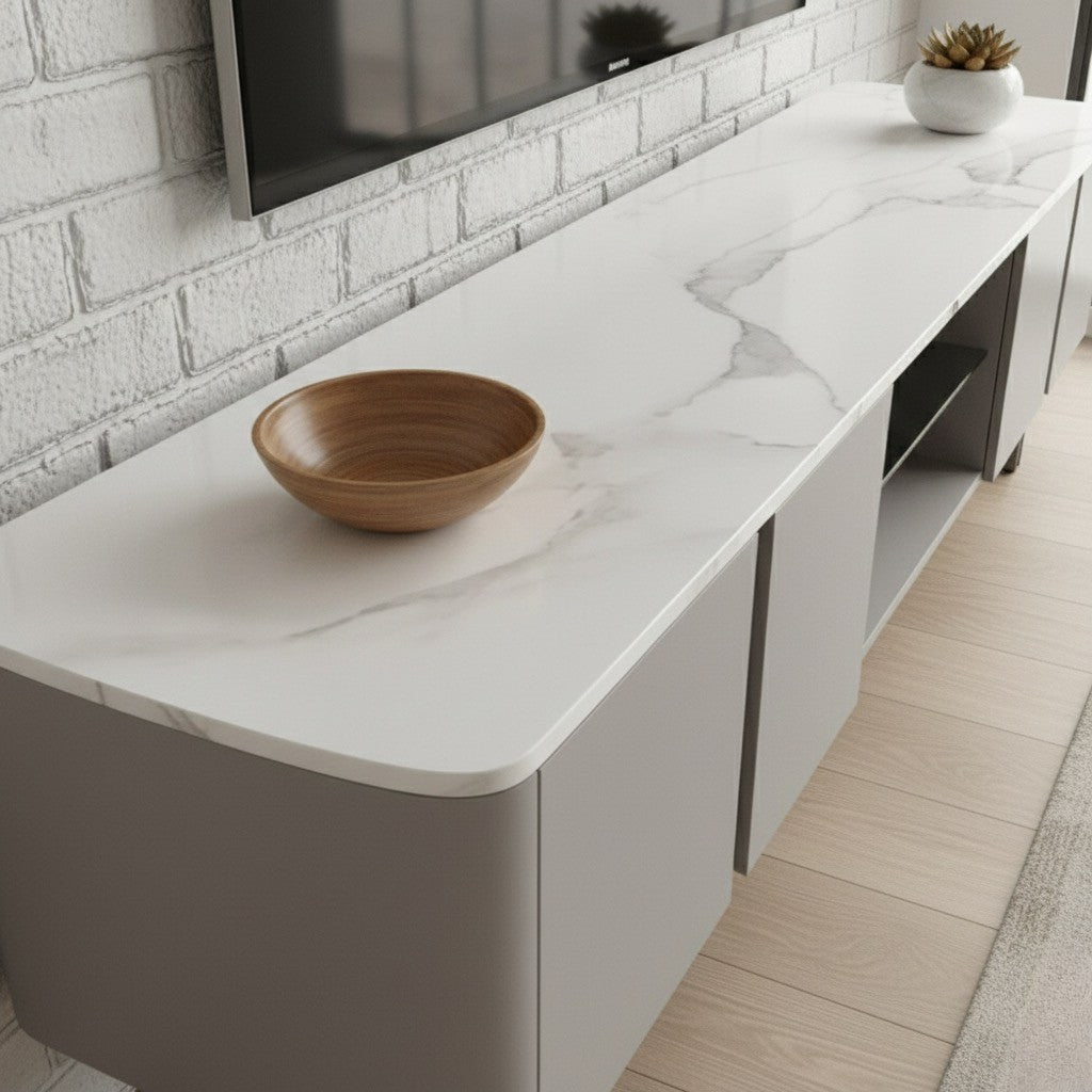 A close-up shot of a modern media console or TV stand. The stand has a white countertop with grey marble-like veining and light grey/taupe base cabinets with rounded edges. On the countertop, there is a round wooden bowl in the foreground and a small potted succulent in a white vase near the back. A flat-screen television is mounted above the console against a white painted brick wall. The floor is light wood with a beige rug visible.